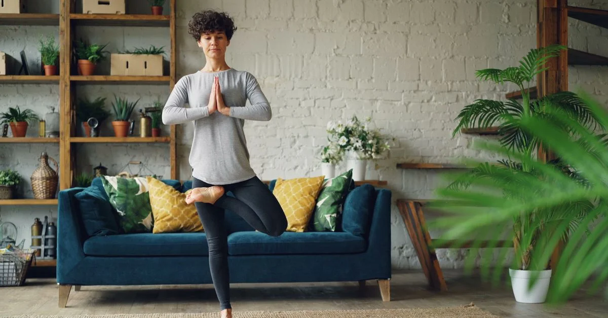 Une femme pratique le yoga dans un salon élégant avec des plantes et un canapé bleu, véhiculant la tranquillité.